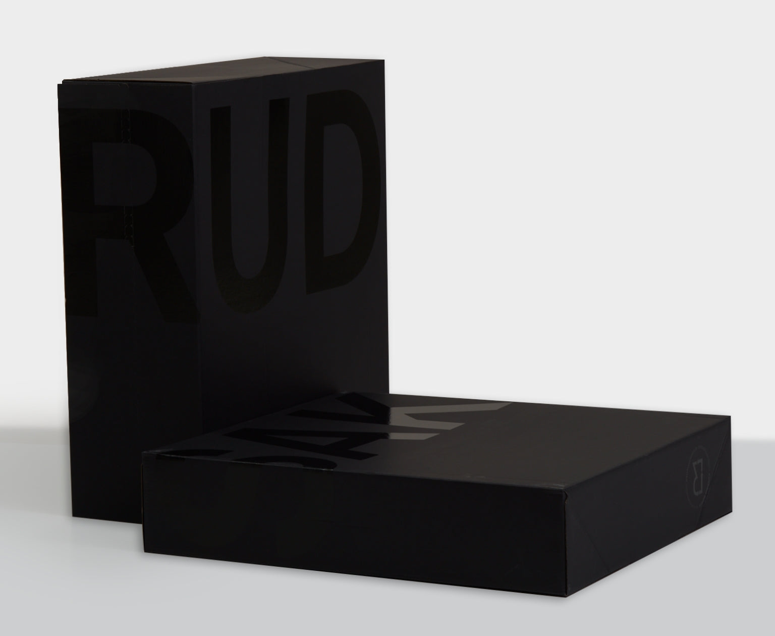 Two sleek, matte black rectangular boxes with subtle glossy black lettering partially spelling RUD and ALK, positioned upright and lying flat against a plain white background.