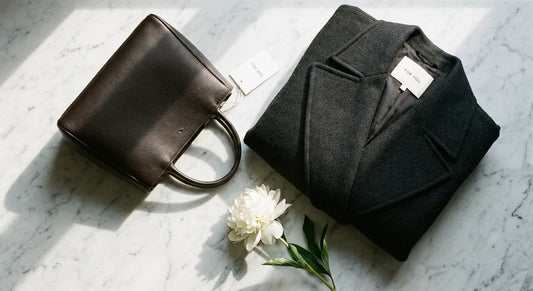 A refined flat lay of a structured dark leather handbag and folded charcoal wool coat beside a single white peony on a marble surface, bathed in soft natural light.