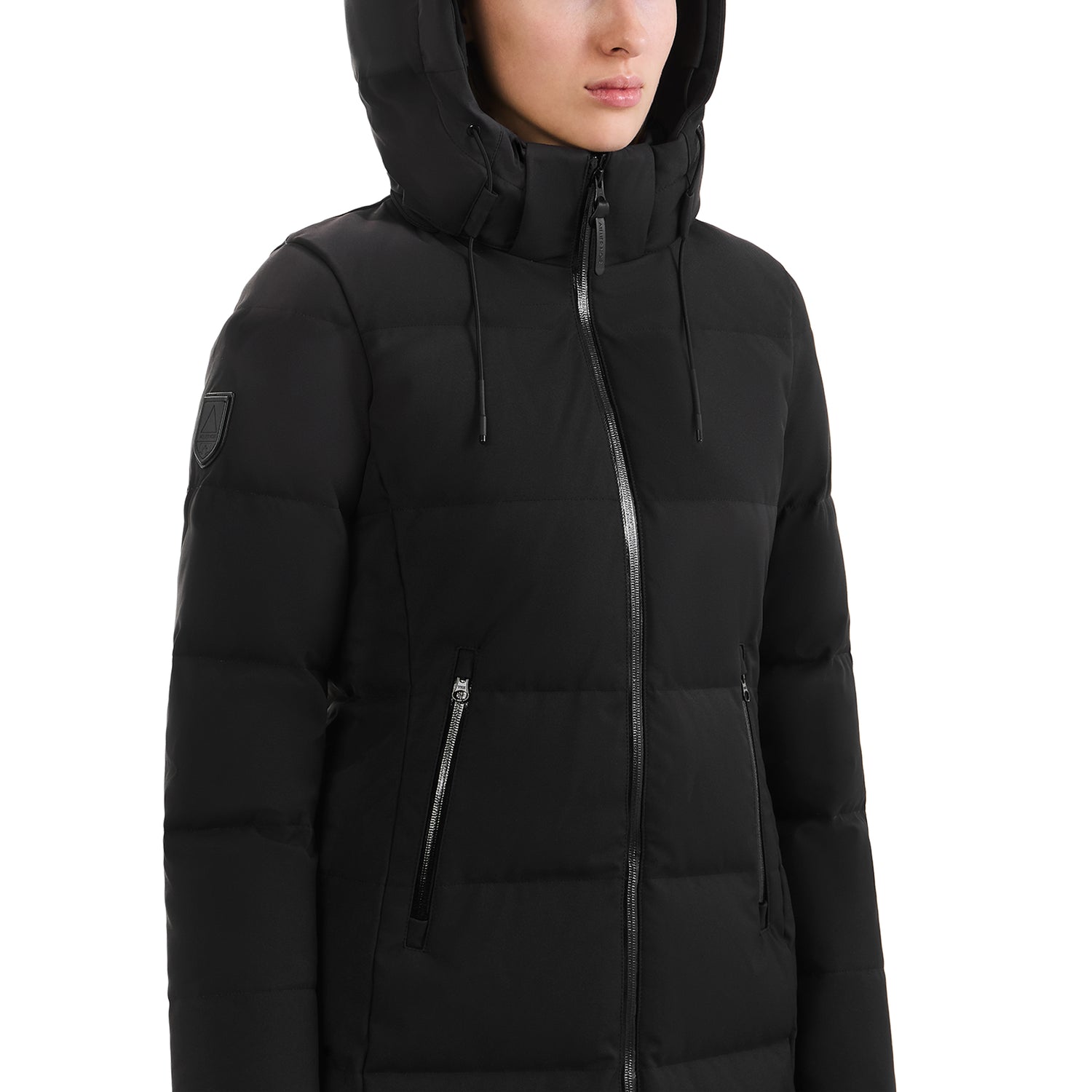 A person wearing the Rudsak SYDNEY COM - 4123511 Black puffer jacket, featuring a hood, wind-resistant fabric, zippered pockets, and a badge on the upper sleeve. The jacket is zipped up and shown from shoulders to mid-thigh as they face slightly sideways.