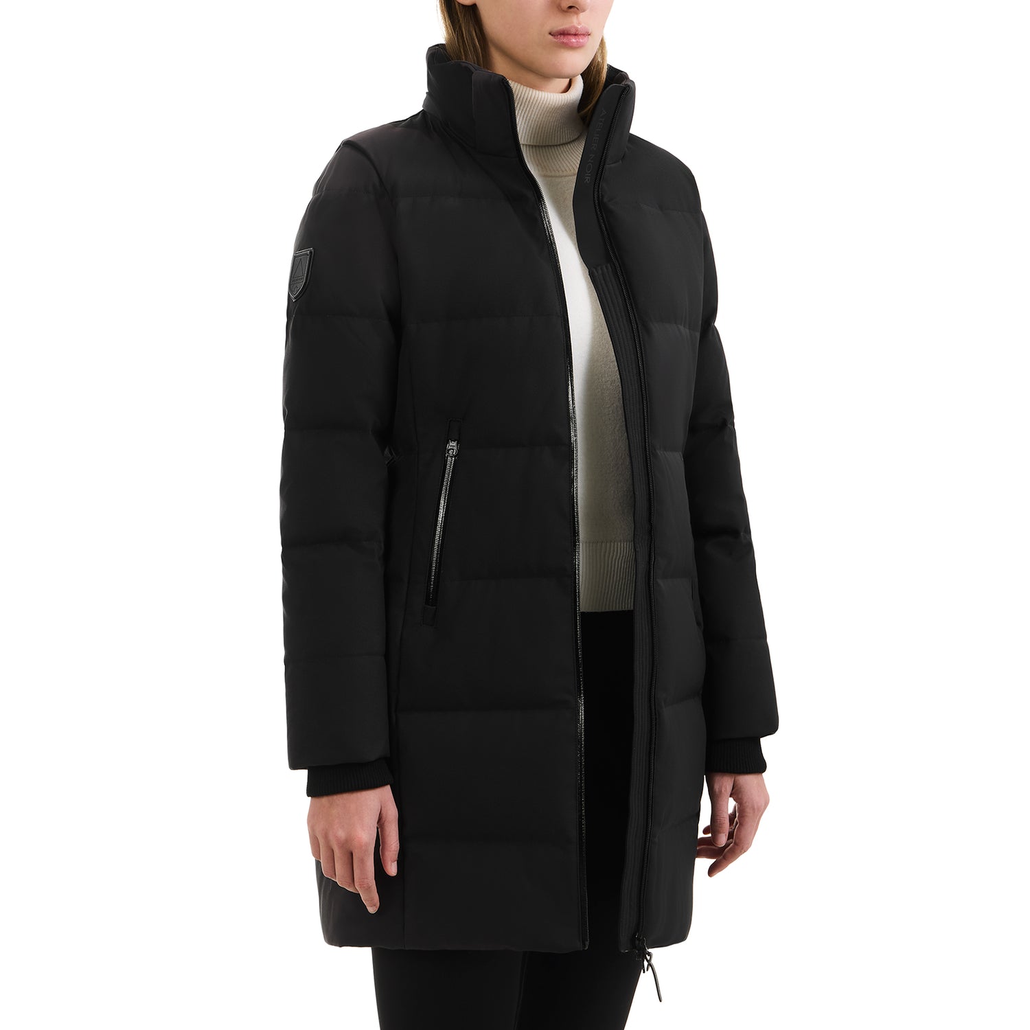 A person wearing the Rudsak SYDNEY COM - 4123511 Black long, quilted, water-repellent winter coat with a high collar and zippered pockets over a light turtleneck and black pants, standing against a white background.