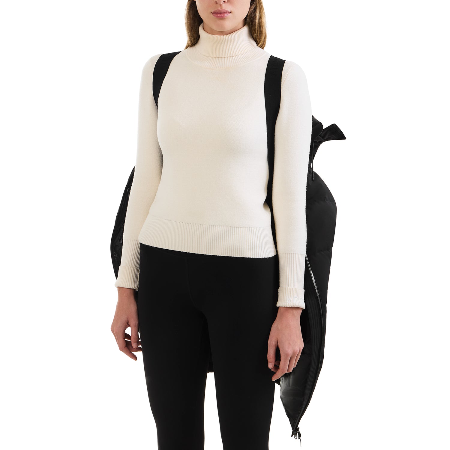 A woman wears a white turtleneck and black pants, carrying the Rudsak SYDNEY COM - 4123511 Black wind-resistant jacket draped over her shoulders like a backpack, while her hands rest relaxed by her sides.