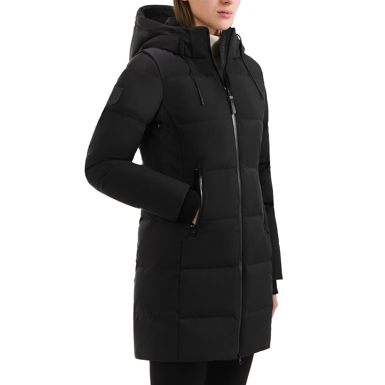 A person wearing the Rudsak SYDNEY COM - 4123511 Black long, quilted, water-repellent winter coat with a hood and zippered pockets stands with one hand in a pocket against a plain white background.