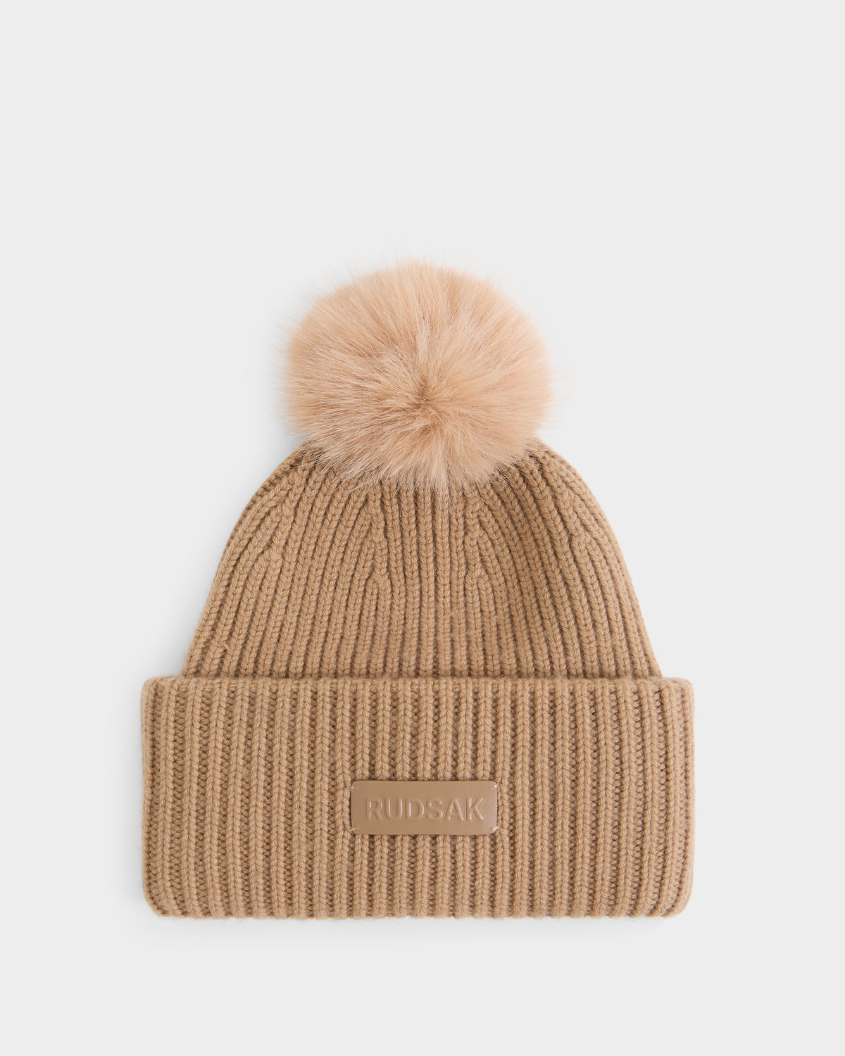 Introducing the DARA - 8425019 Camel by Rudsak, a women’s cashmere tuque in tan with ribbed knit construction, a folded brim, signature RUDSAK patch on the front, and topped with a large, fluffy faux fur pom pom—shown against a plain white background.