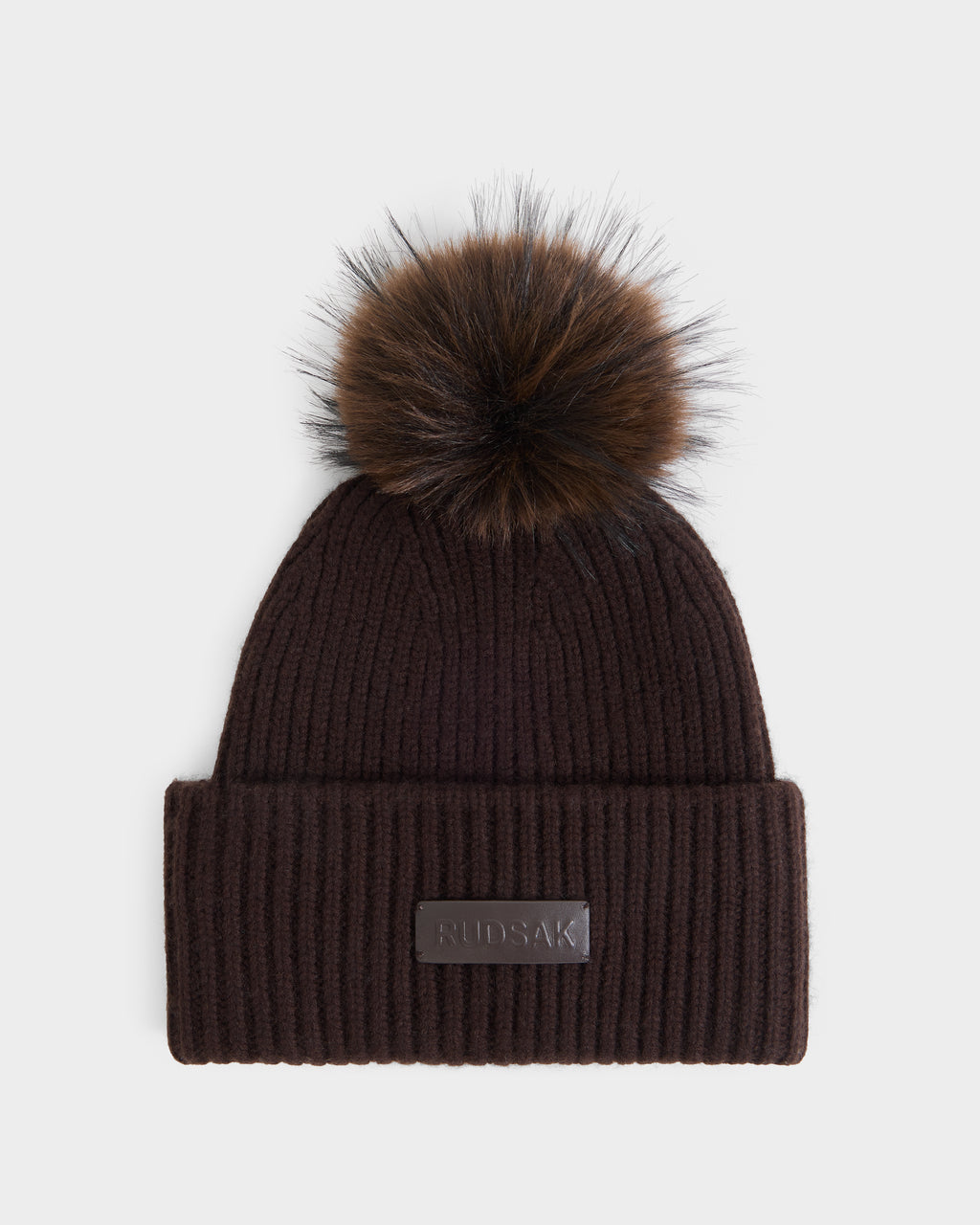 The Rudsak DARA - 8425019 Espresso is a women’s winter beanie in dark brown ribbed knit, featuring a large brown faux fur pom-pom on top and a folded brim accented with a rectangular leather patch embossed with text.