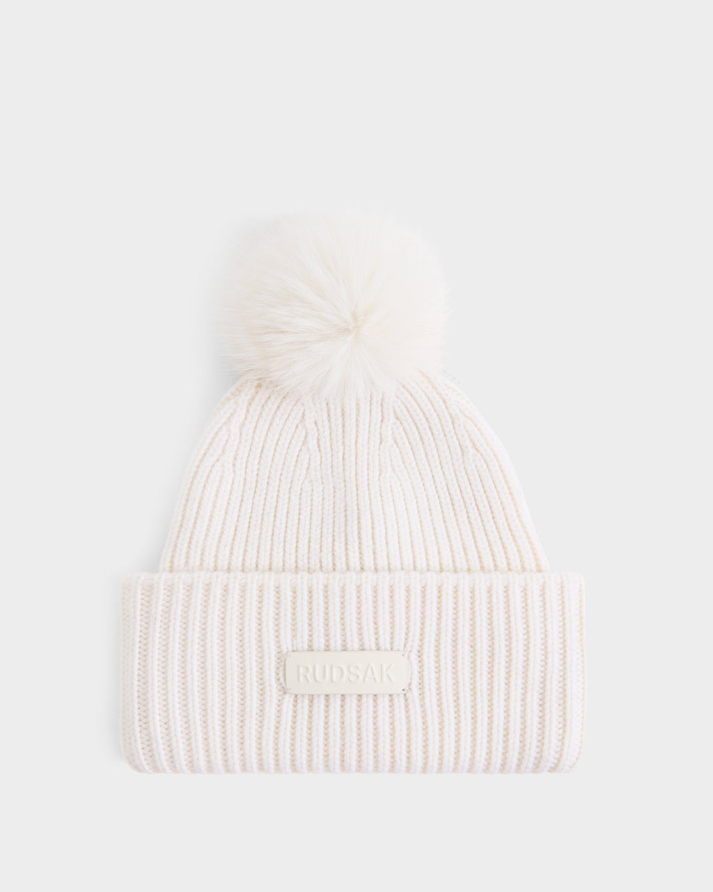 The DARA - 8425019 Vanilla by Rudsak is an off-white ribbed knit women’s tuque featuring a folded brim, a fluffy faux fur pom pom on top, and a signature RUDSAK label on the front. Crafted from a luxurious cashmere wool blend, this hat is shown against a plain white background.