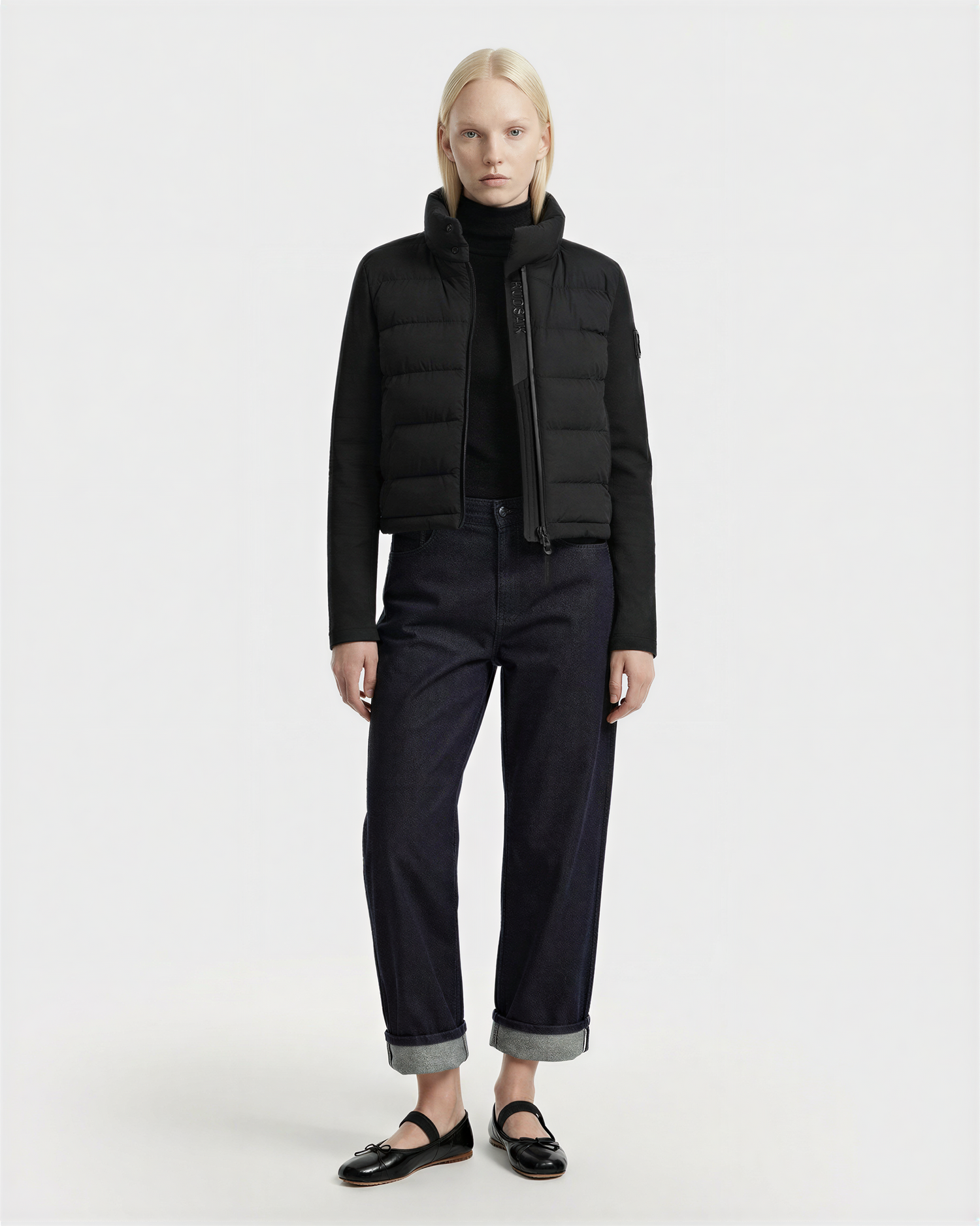 A person with straight blonde hair wears Rudsak’s AUSHA - 8125026 Black women’s cropped down jacket, paired with a black turtleneck, dark cuffed jeans, and black loafers, standing against a plain white background.
