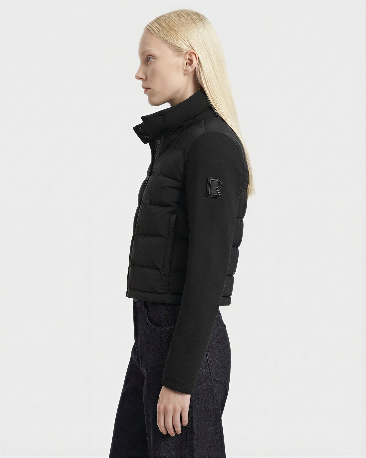 A person with straight blonde hair, wearing the Rudsak AUSHA - 8125026 Black puffer jacket and dark pants, is shown in profile against a plain white background.