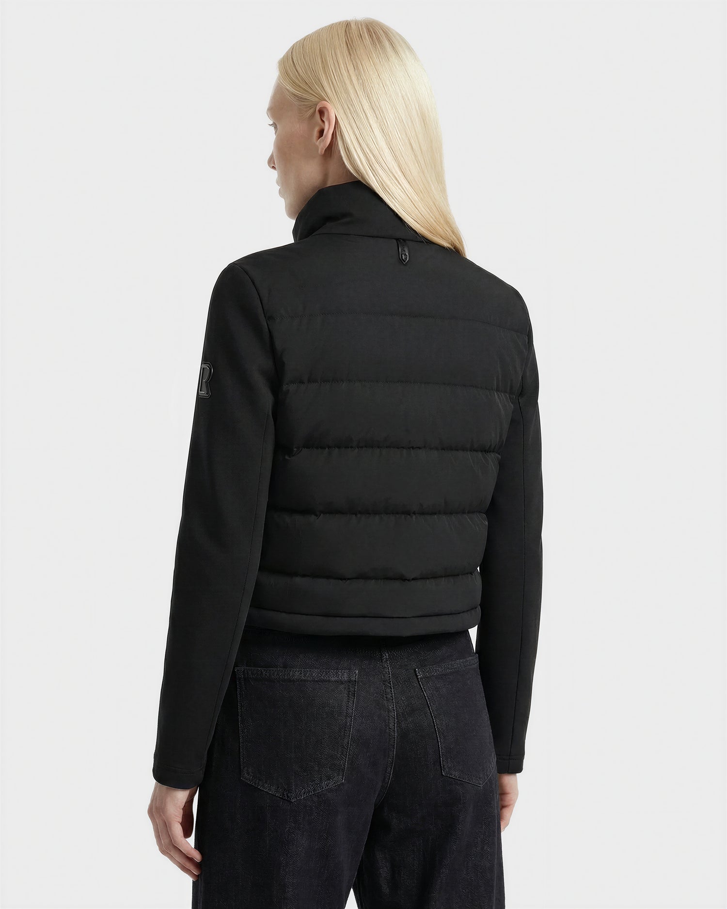 A person with long, straight blonde hair is shown from the back, wearing the Rudsak AUSHA - 8125026 Black women's cropped down jacket and dark jeans against a plain light gray background.