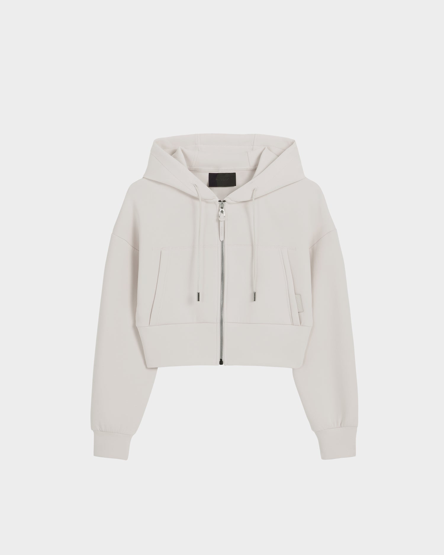 The Rudsak ALAYA - 8525038 Vanilla is a women’s cropped zip-up hoodie in light beige, made from premium double jersey with a drawstring hood, long sleeves, two large front pockets, and a front zipper. Shown on a plain white background.