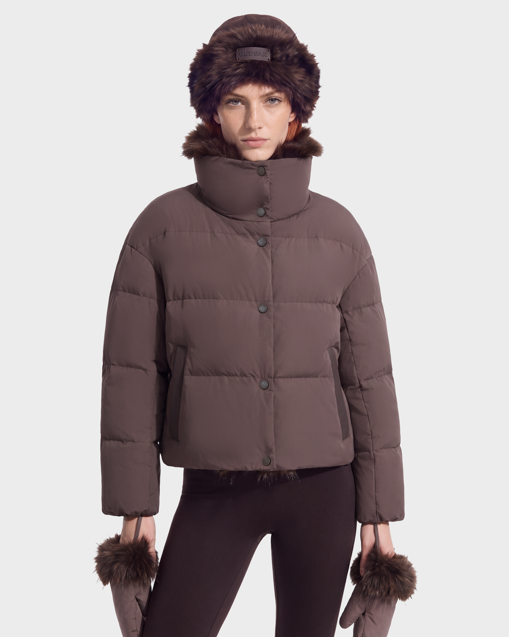 A person wears the Rudsak ALONDRA - 8126992 Espresso brown cropped puffer jacket with a high collar, matching leggings, a faux fur hat, and matching mittens as cozy winter outerwear.