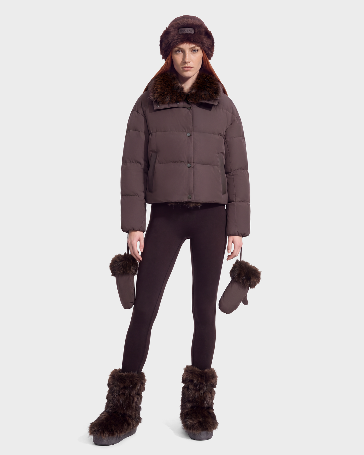 A woman wearing an ALONDRA - 8126992 Espresso cropped puffer jacket with a faux-fur collar, matching leggings, a faux-fur hat, faux-fur boots, and carrying a pair of faux-fur trimmed mittens.