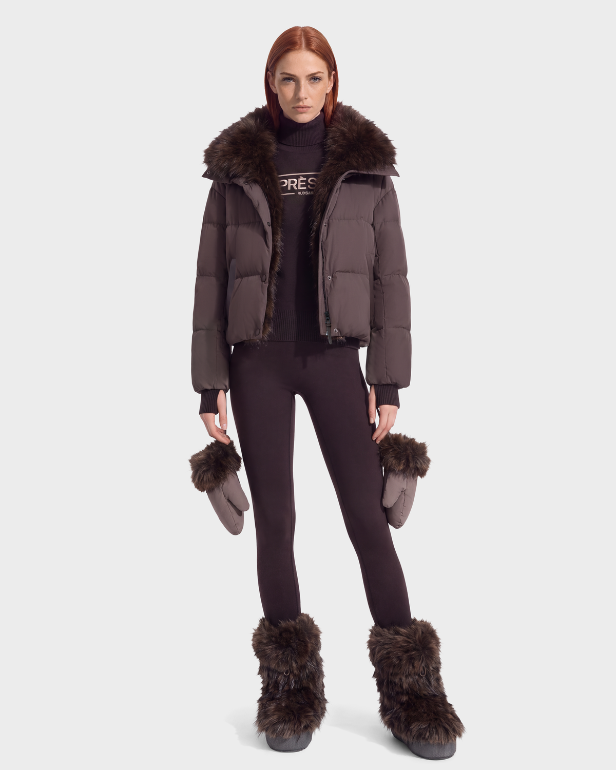 A person wearing an ALONDRA - 8126992 Espresso cropped puffer jacket with a faux fur collar, dark turtleneck, matching leggings, faux fur-trimmed mittens, and faux fur-lined boots.