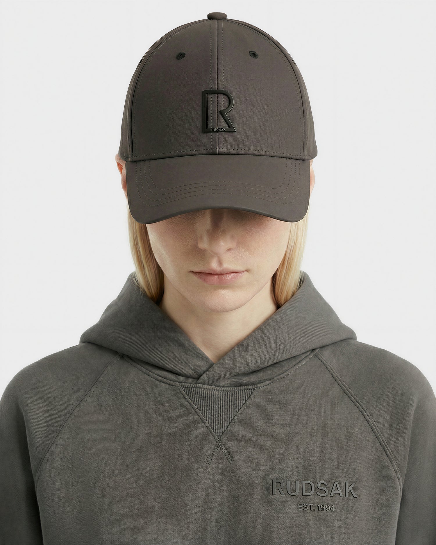 A person wears the Rudsak ARSENE - 9422504 Grey cap with a raised R logo and a matching gray sweatshirt featuring "RUDSAK EST. 1994" embroidery, facing forward with head slightly down.