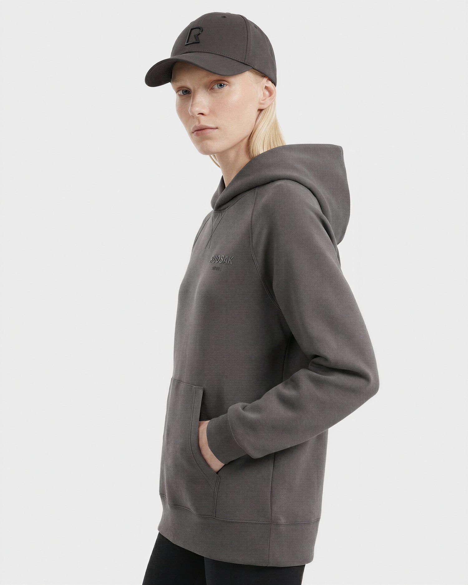 A person with long blond hair wears a dark gray ARSENE - 9422504 Grey hoodie from Rudsak and a matching unisex baseball cap, standing against a white background, facing slightly to the side with one hand in the hoodie pocket.
