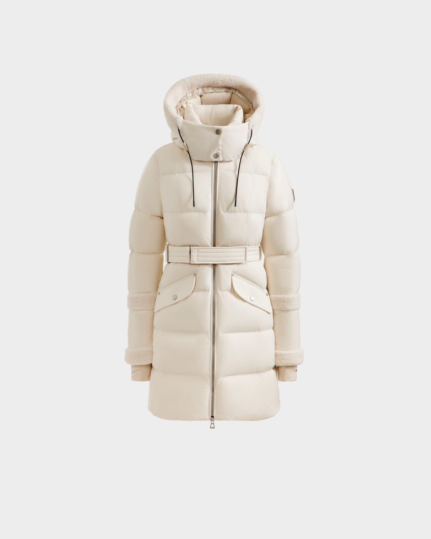 The Rudsak AUDREY L - 8124585 Vanilla is a cream knee-length down puffer jacket with a hood, high collar, belted waist, front zipper, and large snap-button flap pockets—made with RDS certified down. Shown on a white background.