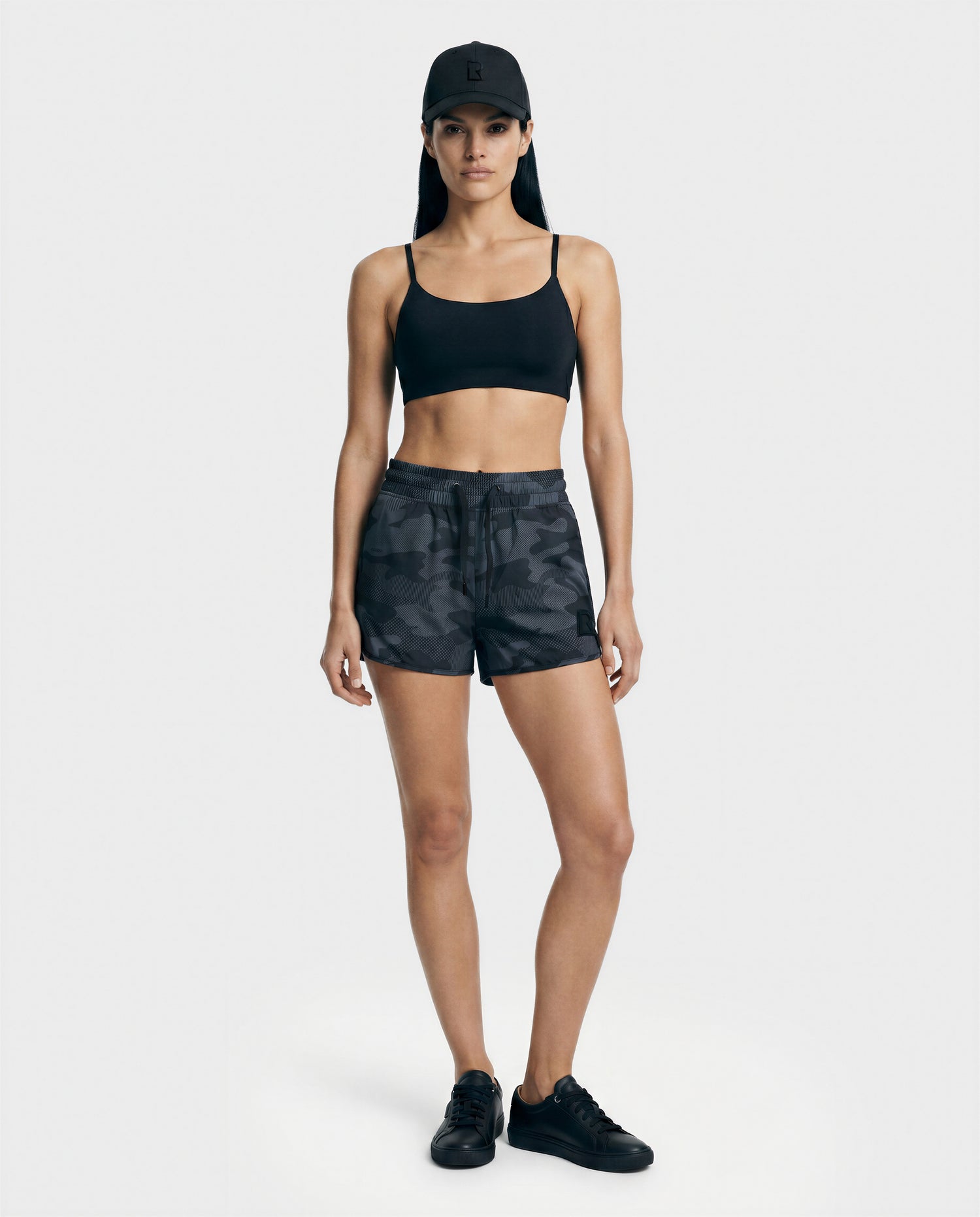 A woman faces forward against a plain light background, wearing the Rudsak CAPRI - L523009 BLACK sports bra, camo shorts, black sneakers, and a black cap.