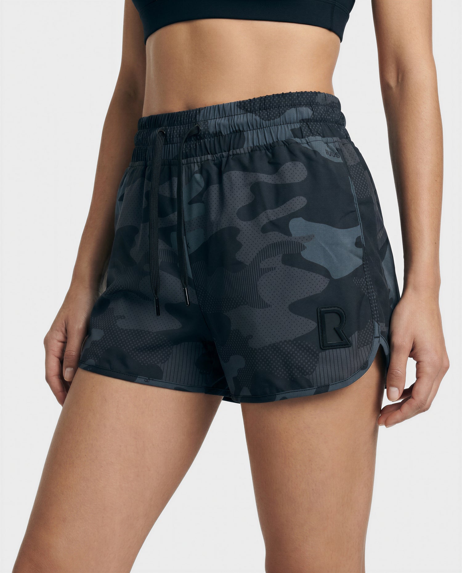 A person in athletic wear featuring Rudsak CAPRI - L523009 BLACK camo shorts with a black R logo on the left leg and a black sports bra, standing with one hand by their side.