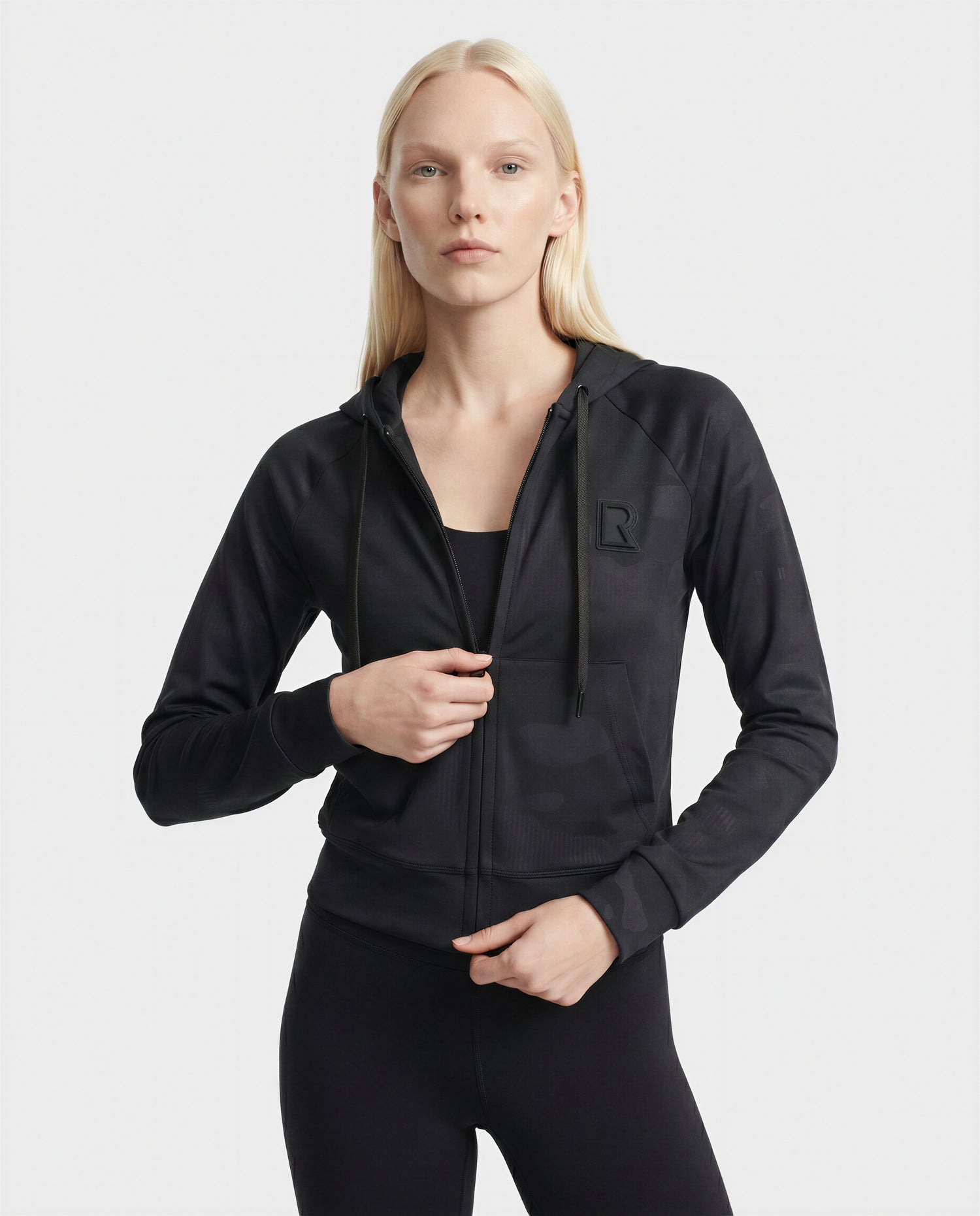 A woman with long blonde hair models the Rudsak SAMMY - L523014T camouflage black cropped hoodie with a raised R logo on the chest and black pants, standing against a plain light background for an effortlessly luxurious look.