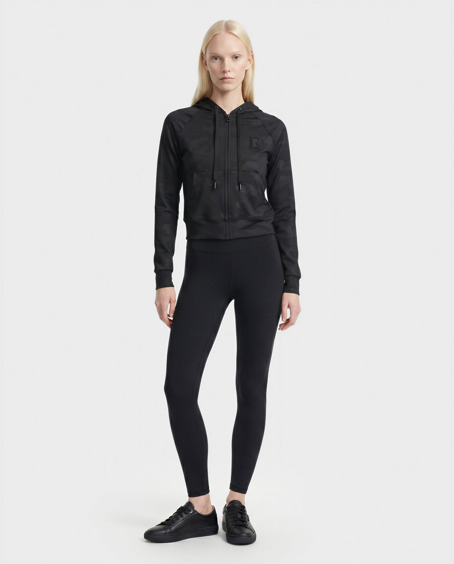 A woman with long blonde hair stands facing forward, wearing the Rudsak SAMMY - L523014T Top in Camouflage Black against a plain white background.