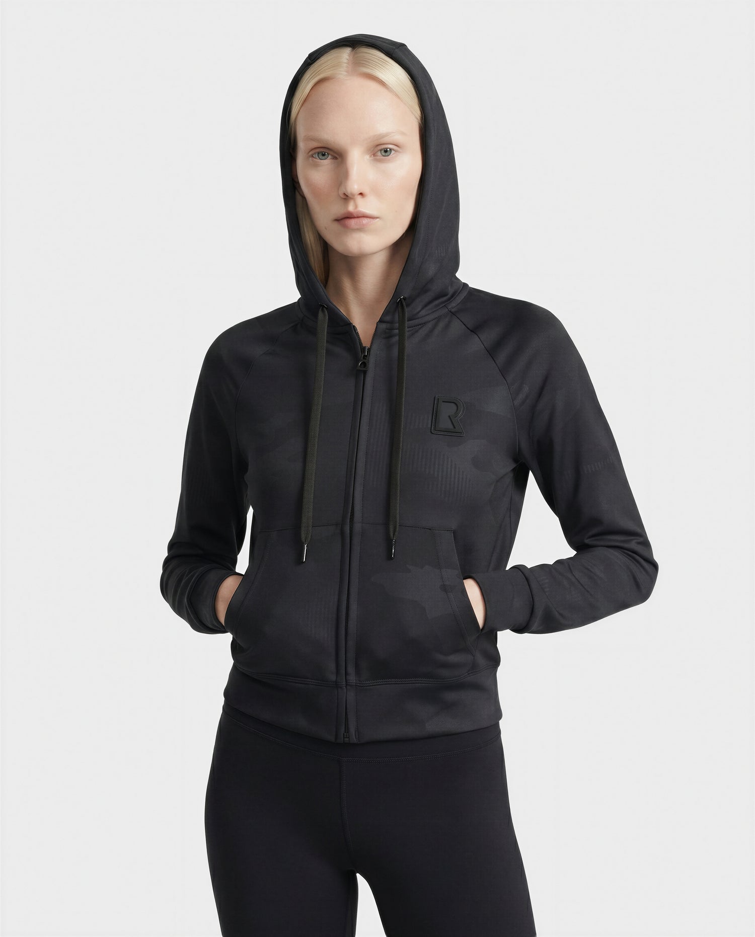 A woman with straight blonde hair wears the Rudsak SAMMY - L523014T Camouflage Black top, a luxury cropped hoodie with subtle camo print and a raised logo, hands resting in the front pockets against a plain light background.
