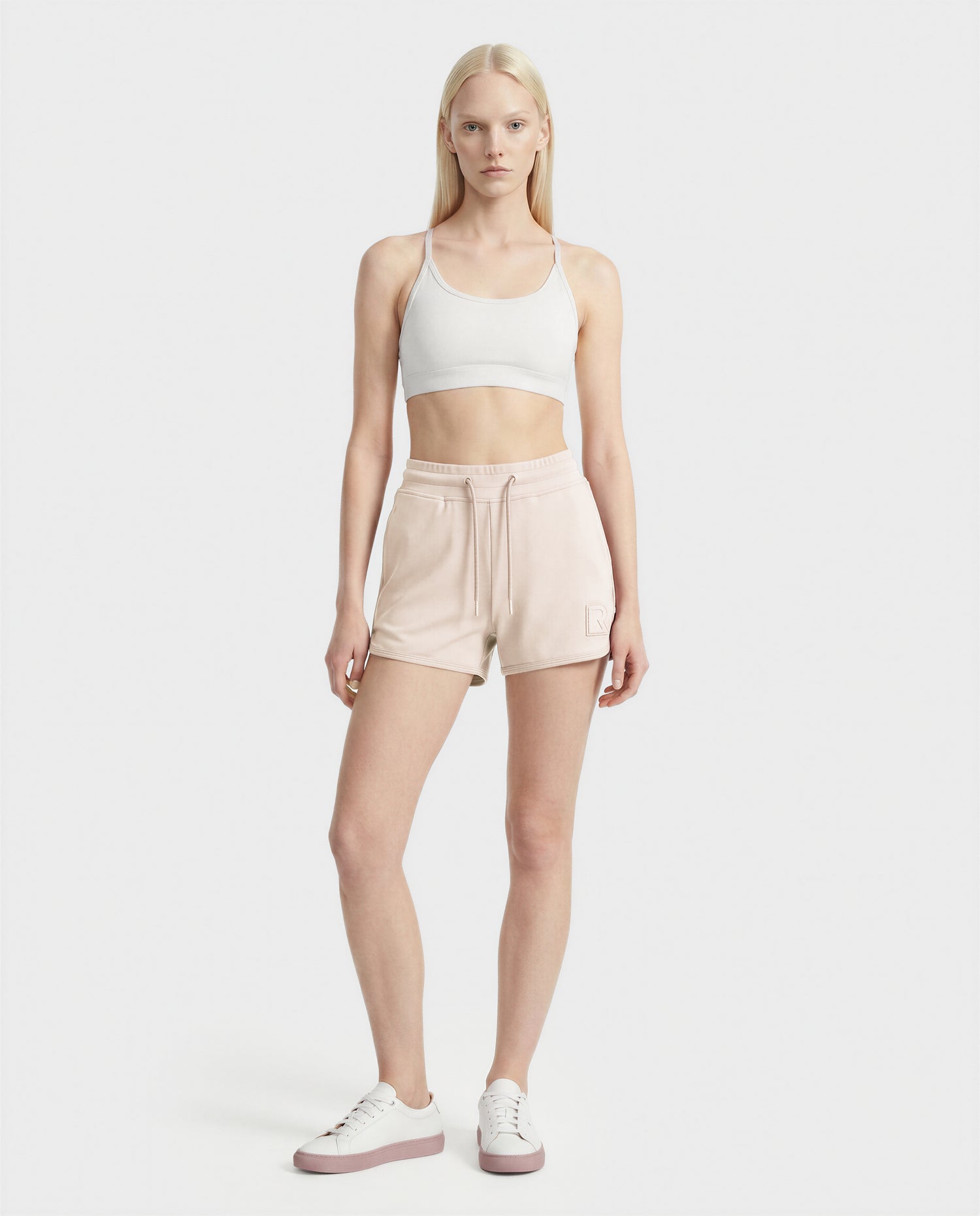 A person with long blonde hair stands against a plain white background, wearing a white sports bra, SAMMY - L523014B cream bottoms by Rudsak, and white sneakers.