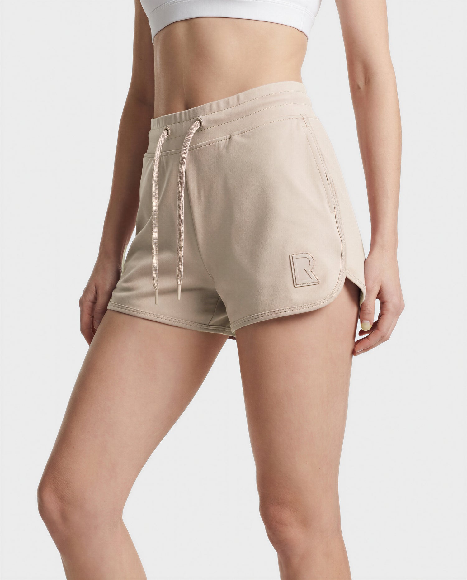 A person wearing Rudsak's SAMMY - L523014B cream shorts with a raised R logo, paired with a white sports bra, poses against a plain white background.