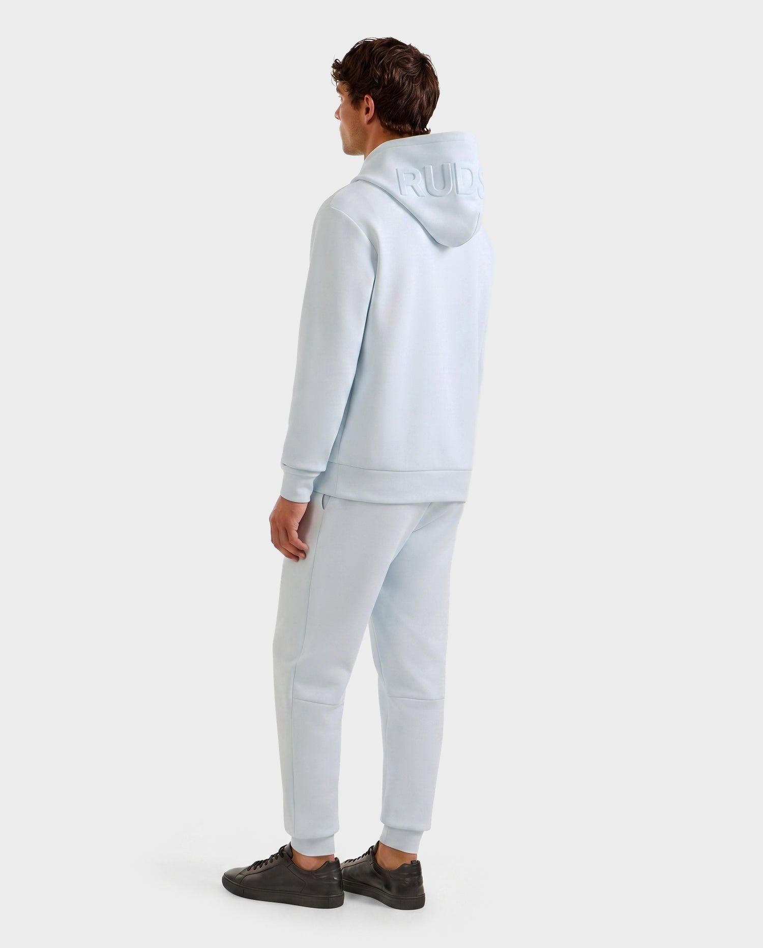 A person faces away, wearing the Rudsak LEROY - U525509 hoodie and matching joggers in sky blue (M). "RUD" lettering appears on the hood. They have short brown hair, dark sneakers, and stand against a plain light background.