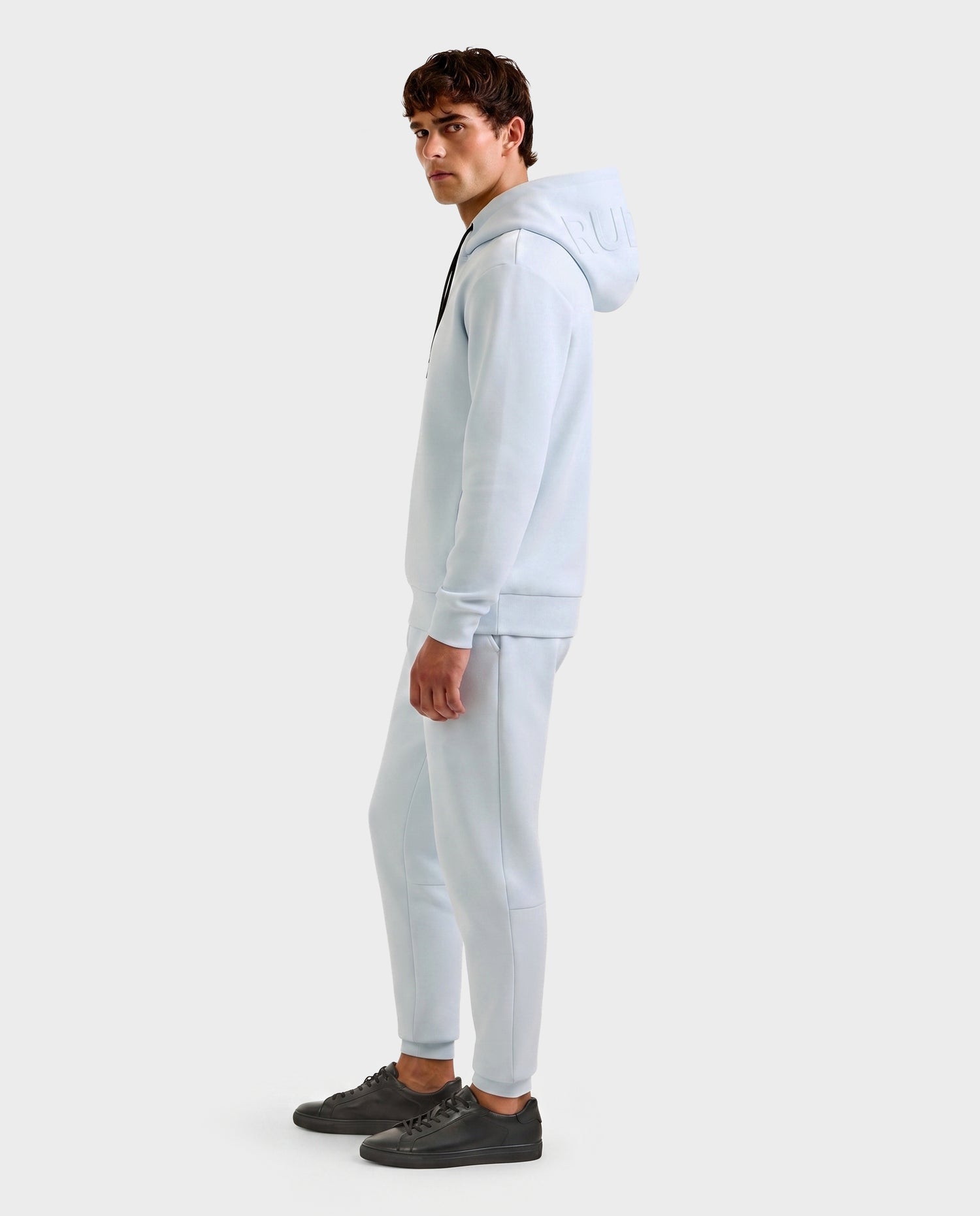 A young man stands sideways in a Rudsak LEROY - U525509 Sky Blue M premium unisex hoodie and matching sweatpants with black sneakers, set against a plain light gray background.