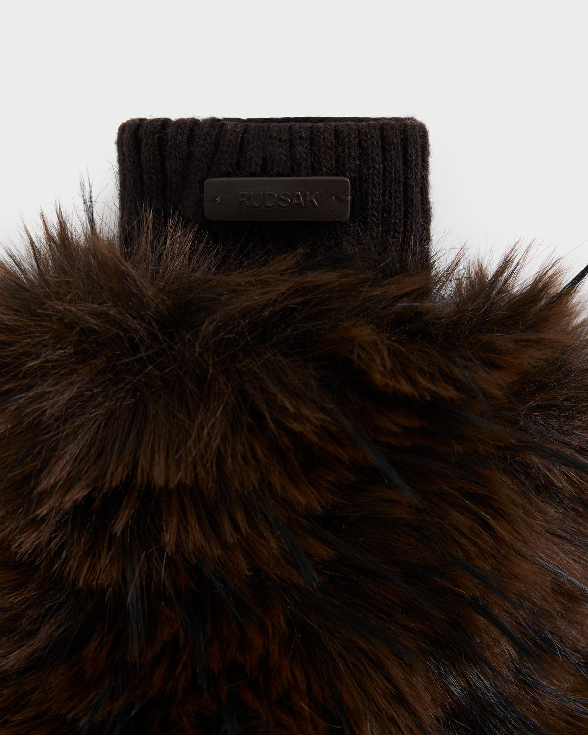 Close-up of the BELLA - 8425006 Mix Espresso mitten by Rudsak, showcasing its brown faux fur exterior with knit material and a black leather tag labeled “RUDSAK” stitched onto the knit section, set against a plain white background. This cozy winter glove is the ideal après-ski accessory.