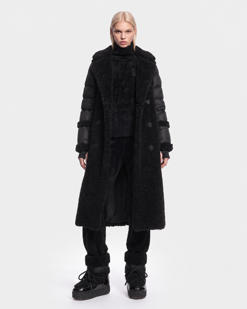 A person with long blonde hair wears the Rudsak NOELLE - 8126971 Black faux fur coat, black pants, and chunky black shoes, standing against a white background—perfect as a winter jacket.
