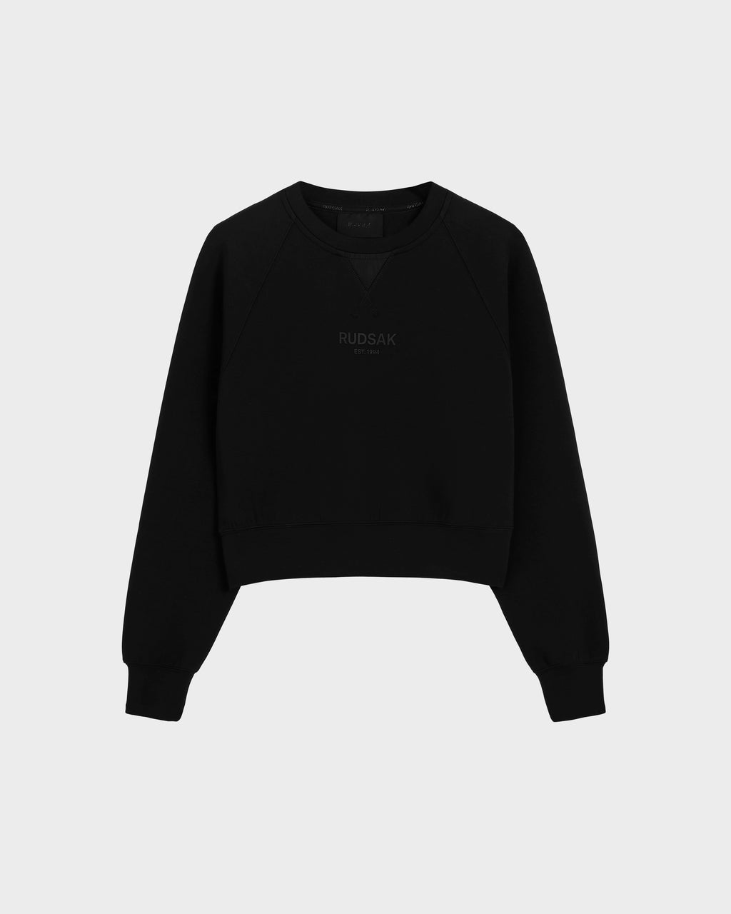 The Rudsak BETSY - 8525087 Black is a black cotton blend crewneck sweater with ribbed cuffs and hem, featuring a subtle RUDSAK print on the chest, shown against a plain light gray background.