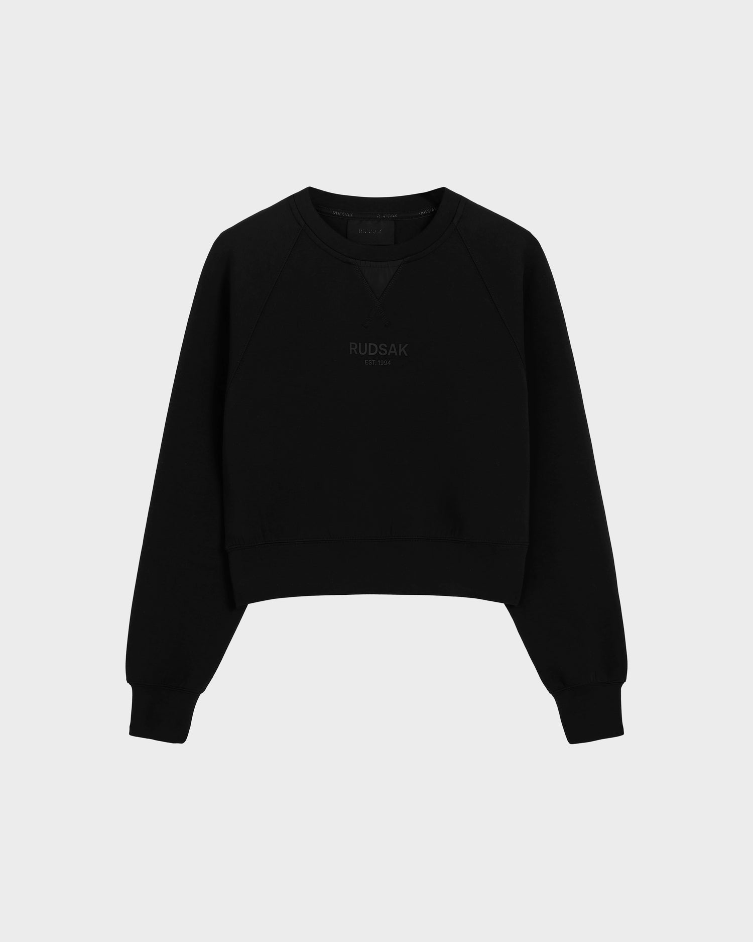 The Rudsak BETSY - 8525087 Black is a black cotton blend crewneck sweater with ribbed cuffs and hem, featuring a subtle RUDSAK print on the chest, shown against a plain light gray background.