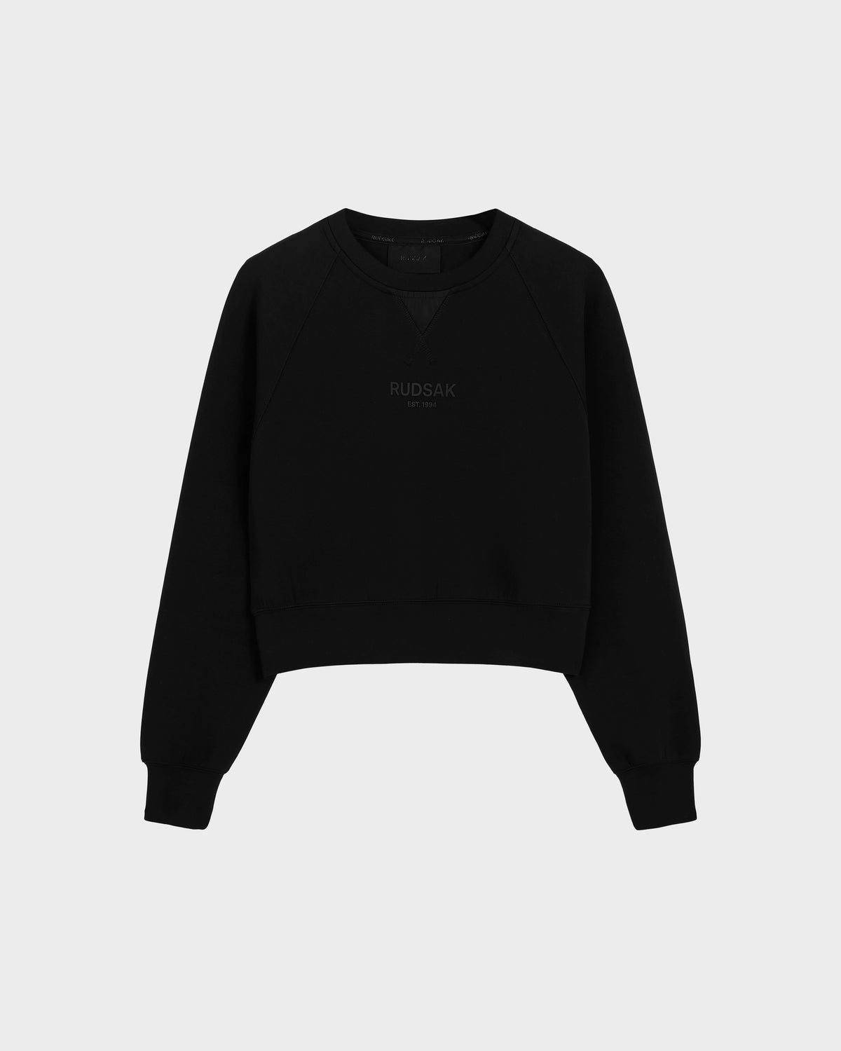 The Rudsak BETSY - 8525087 Black is a black cotton blend crewneck sweater with ribbed cuffs and hem, featuring a subtle RUDSAK print on the chest, shown against a plain light gray background.