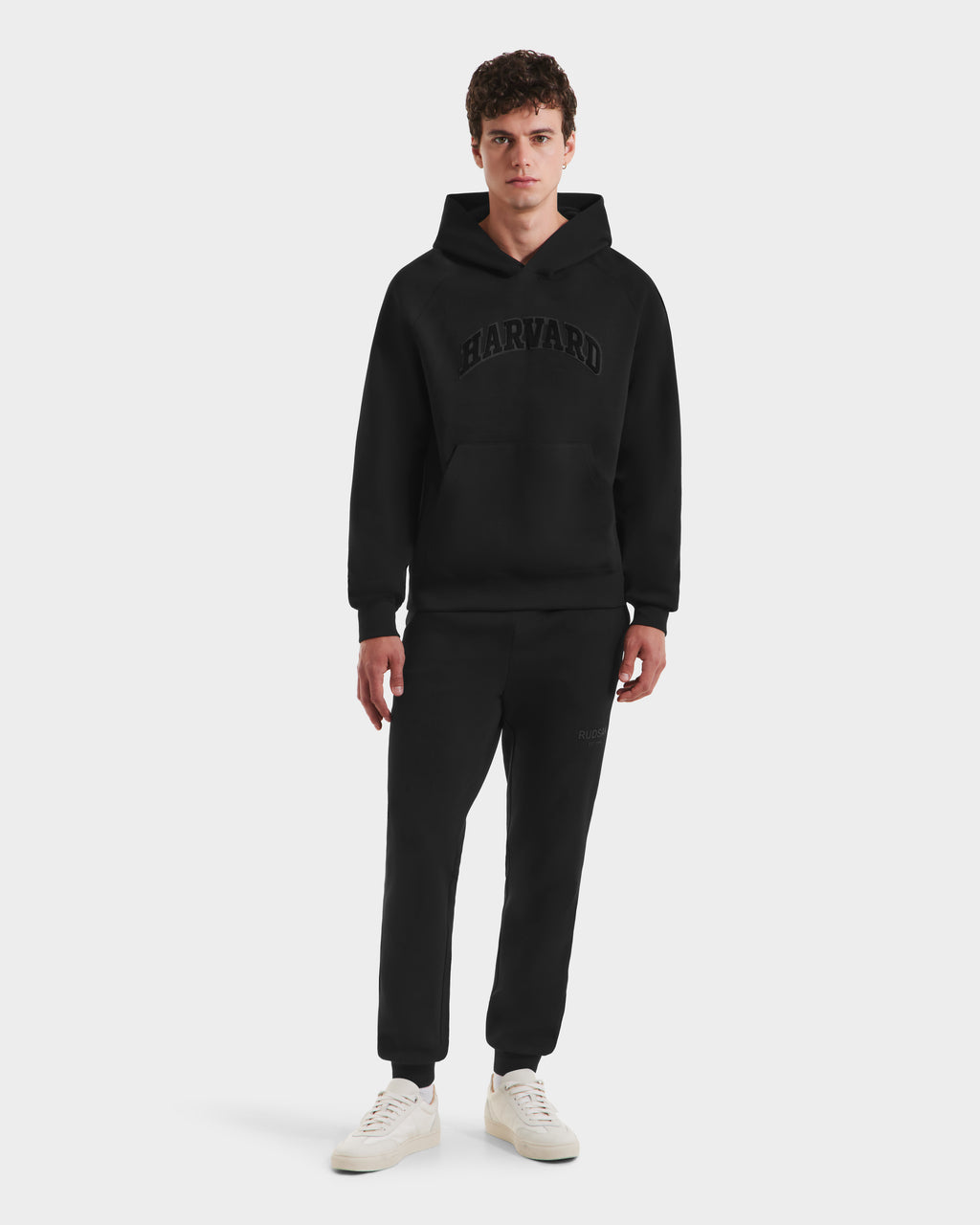 A young man faces forward in a plain white setting, wearing Rudsak's BRIAR B HV - H525509 Black hoodie featuring bold chest text, paired with black sweatpants and white sneakers.