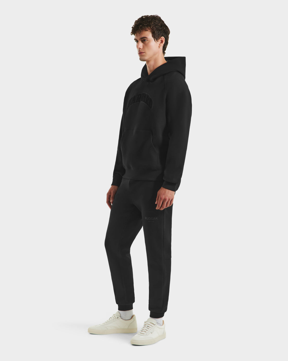 A person stands against a plain white background wearing Rudsak’s limited-edition BRIAR B HV - H525509 Black hooded sweatshirt and matching jogger pants with white sneakers.