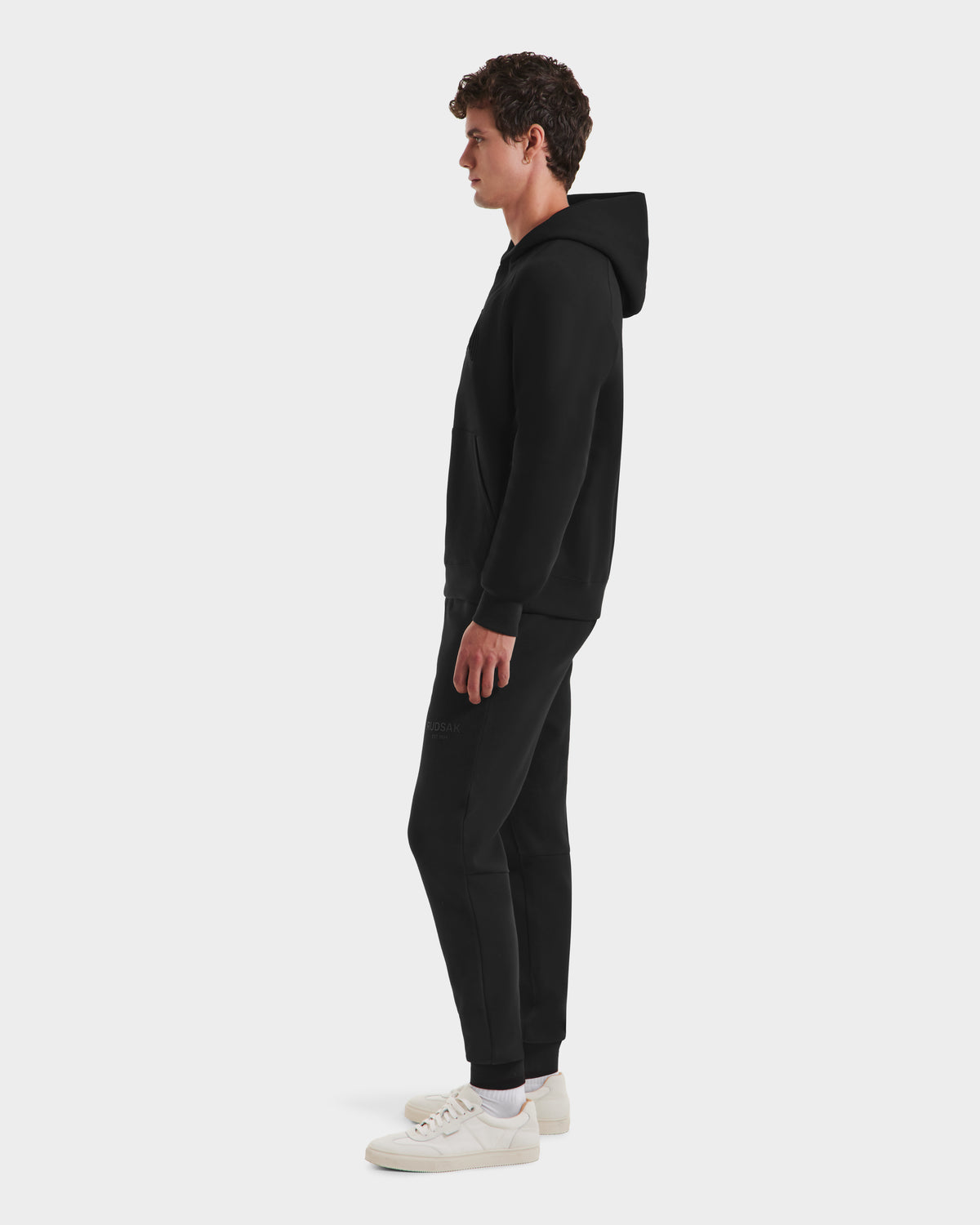A man stands in profile against a plain background, wearing the limited-edition Rudsak BRIAR B HV - H525509 Black Harvard hoodie, paired with black jogger pants and white sneakers.