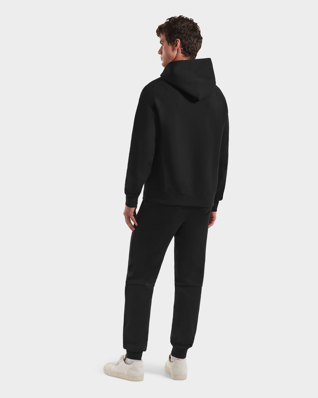 A person stands facing away, wearing a Rudsak BRIAR B HV - H525509 Black hoodie and matching black sweatpants with white sneakers, set against a plain white background.