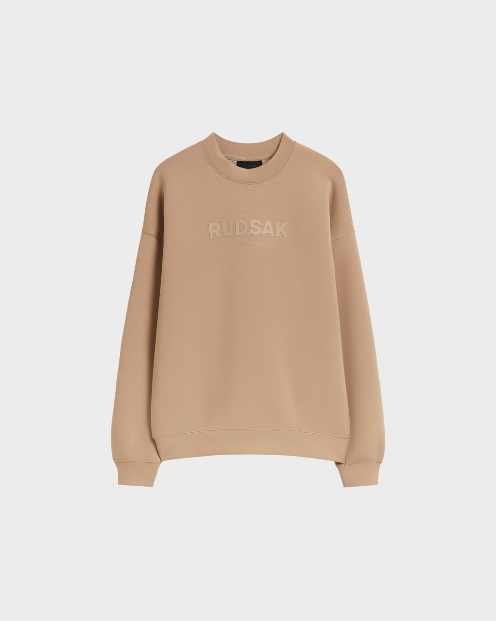 The Rudsak BRYN - 9524007 Almond W is a beige cotton crewneck sweatshirt with long sleeves and genderless sizing, featuring "RUDSAK" embroidered on the front and shown against a plain white background.