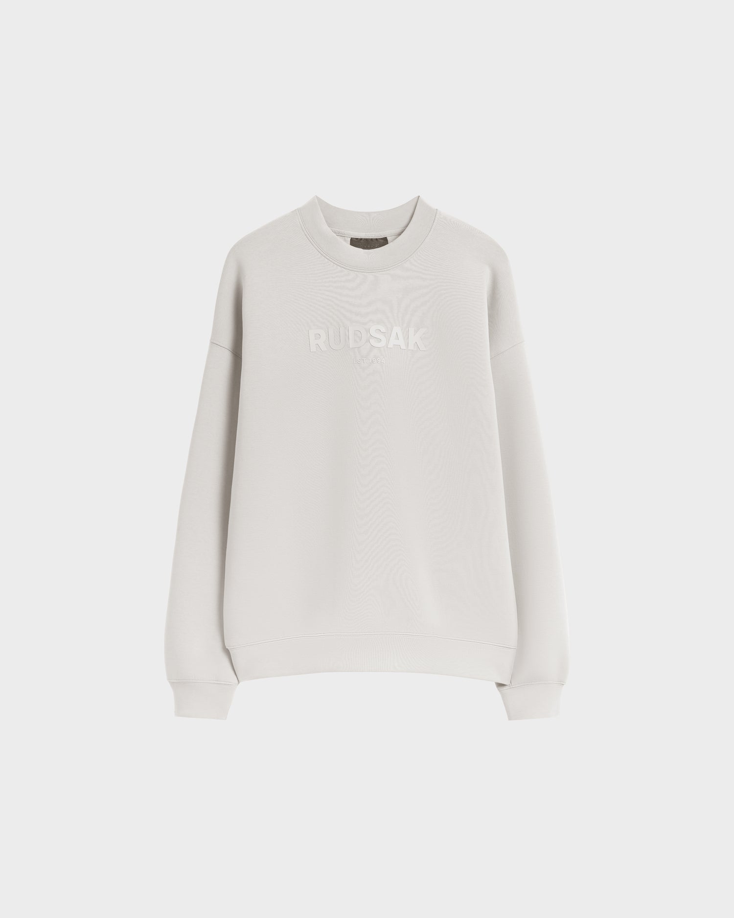 The BRYN - 9524007 Vanilla M by Rudsak is a light grey, long-sleeve cotton crewneck sweatshirt with white RUDSAK embroidery on the front, shown on a white background. Designed for an effortless everyday fit with genderless sizing.