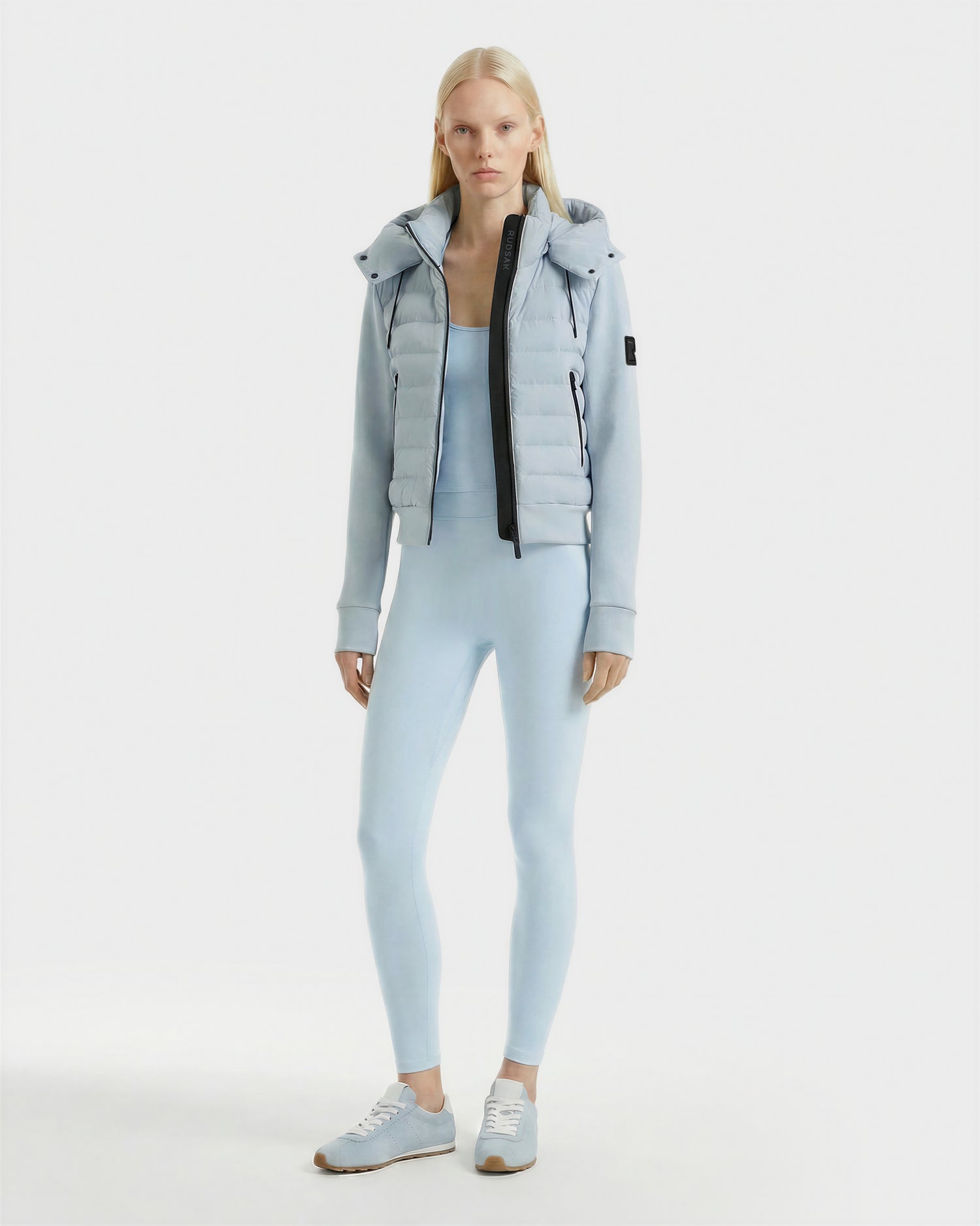 A woman with long blonde hair wears the Rudsak CASSA - 8125057 Powder Blue jacket over matching light blue activewear and sneakers, standing against a plain white background.