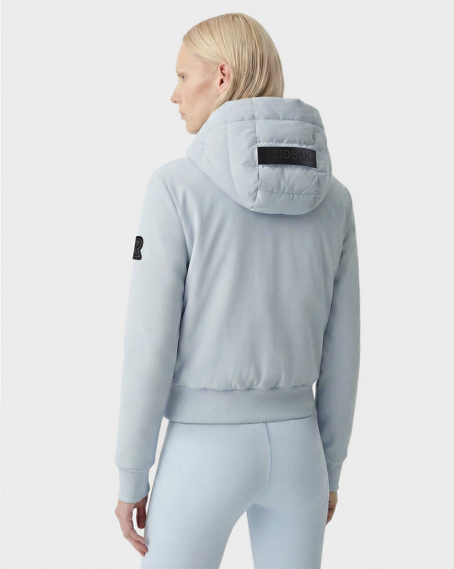 A person with straight blonde hair is seen from the back wearing the Rudsak CASSA - 8125057 Powder Blue women's transitional jacket and matching leggings. The lightweight cropped jacket features a black arm patch, logo hood, and a plain white background.