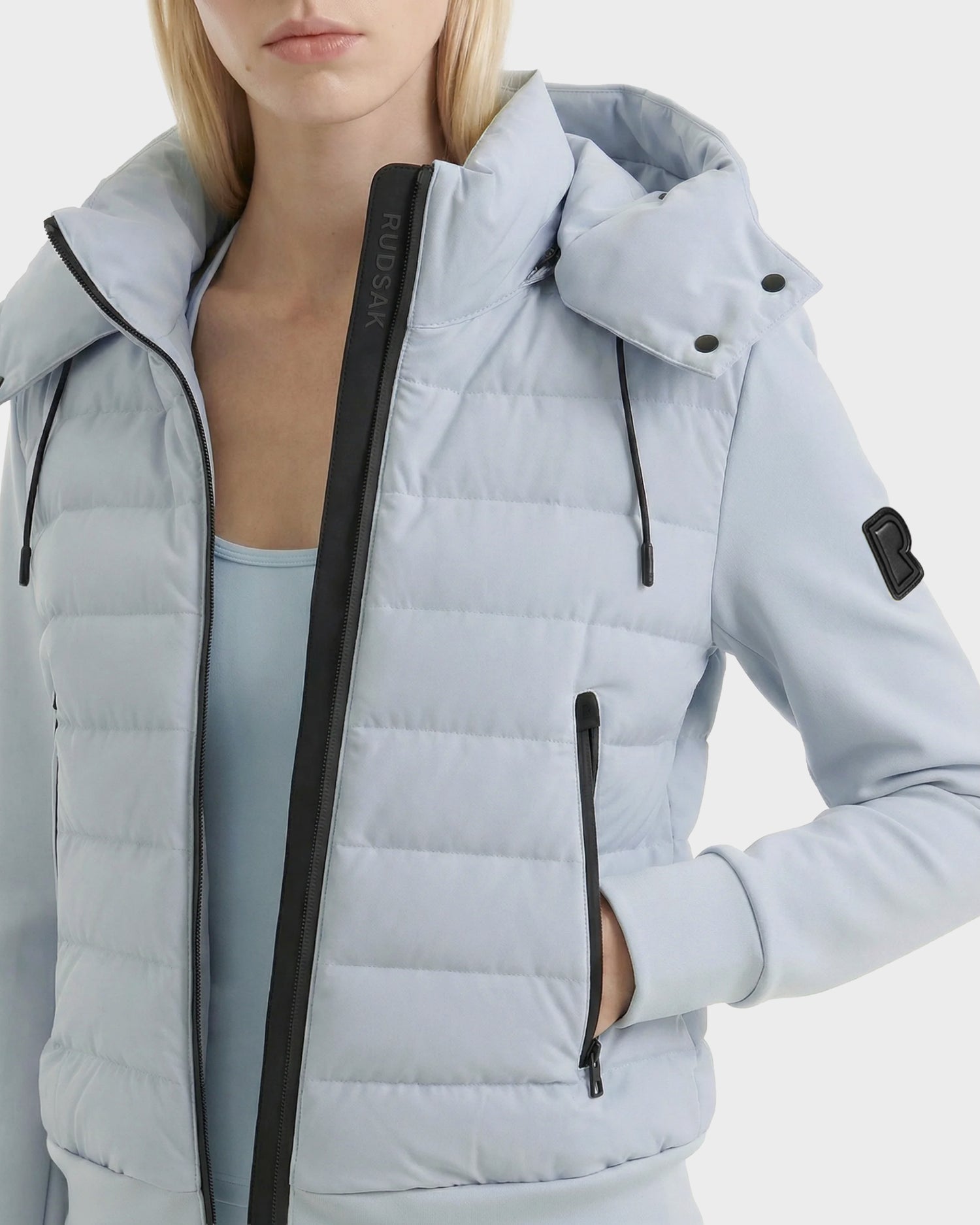A woman models the Rudsak CASSA - 8125057 Powder Blue women's transitional jacket with black zippers, a sleeve logo patch, and her left hand in the zippered side pocket, shown cropped from mid-torso up against a white background.