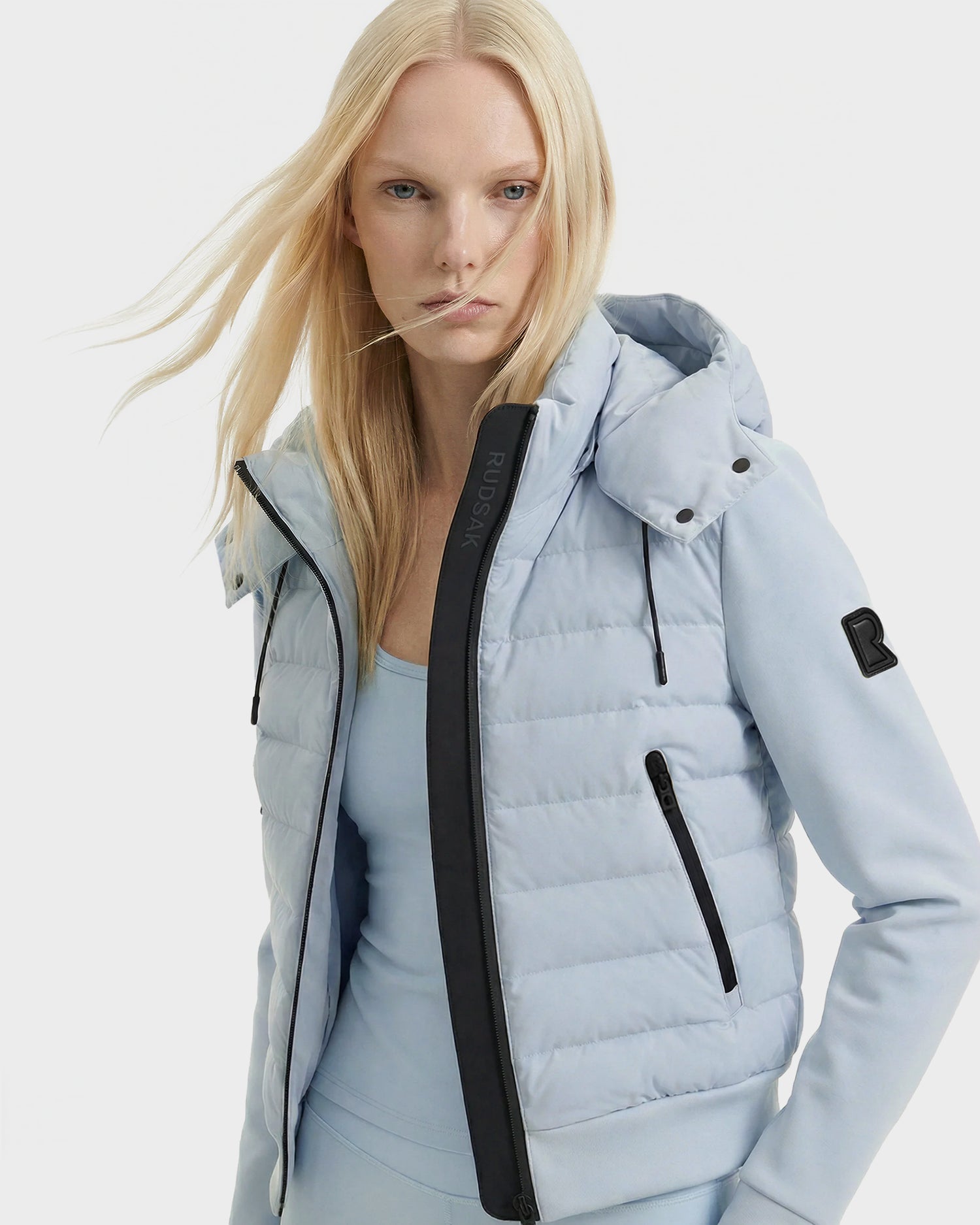 A person with long, straight blonde hair wears the Rudsak CASSA - 8125057 Powder Blue lightweight cropped jacket with a hood and black zipper, layered over a matching top—an ideal choice for transitional weather.