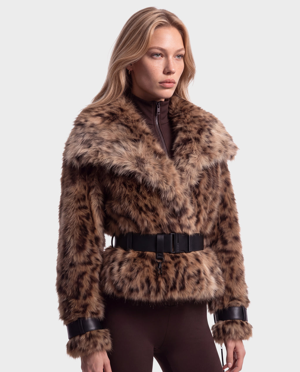 A person wearing a Rudsak CHLOE - 8125619 Leopard short, faux fur jacket with a black waist belt over a brown high-neck top.