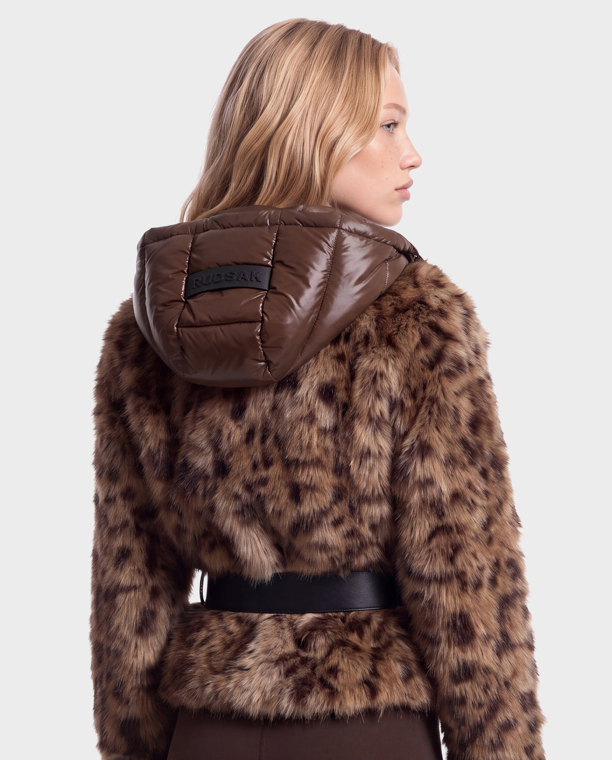 A person wearing a Rudsak CHLOE - 8125619 Leopard faux fur jacket with a brown hooded puffer collar and a black belt.