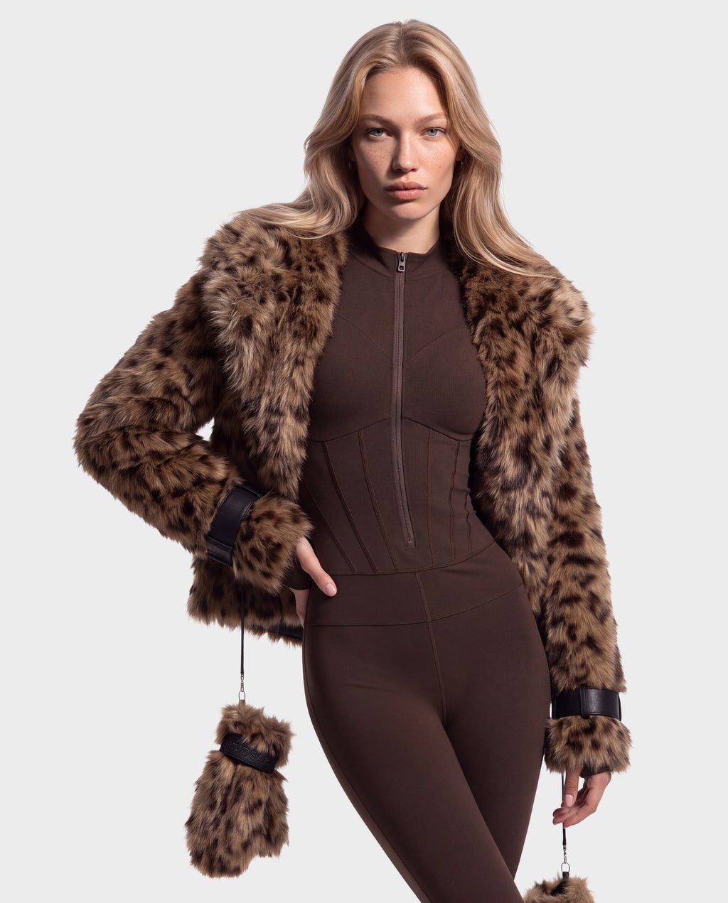 A person wearing a brown zip-up jumpsuit and a Rudsak CHLOE - 8125619 Leopard faux-fur jacket with matching mittens attached to the cuffs, perfect for a winter wardrobe.
