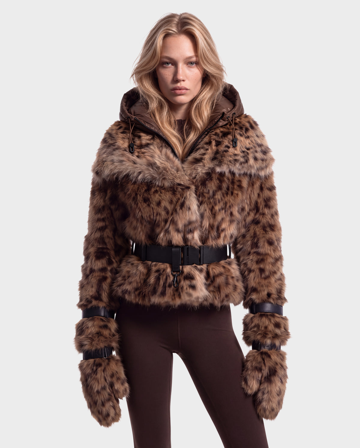 A person wearing a Rudsak CHLOE - 8125619 Leopard faux fur jacket with a black buckled belt, matching leopard-print mittens, and brown leggings.