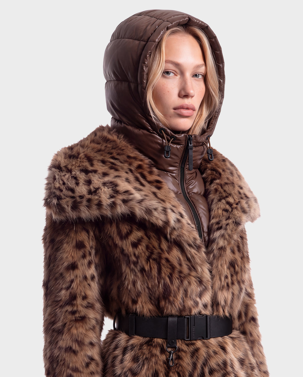 A woman wears a brown puffer hood over a Rudsak CHLOE - 8125619 Leopard faux fur jacket cinched with a black buckled belt.