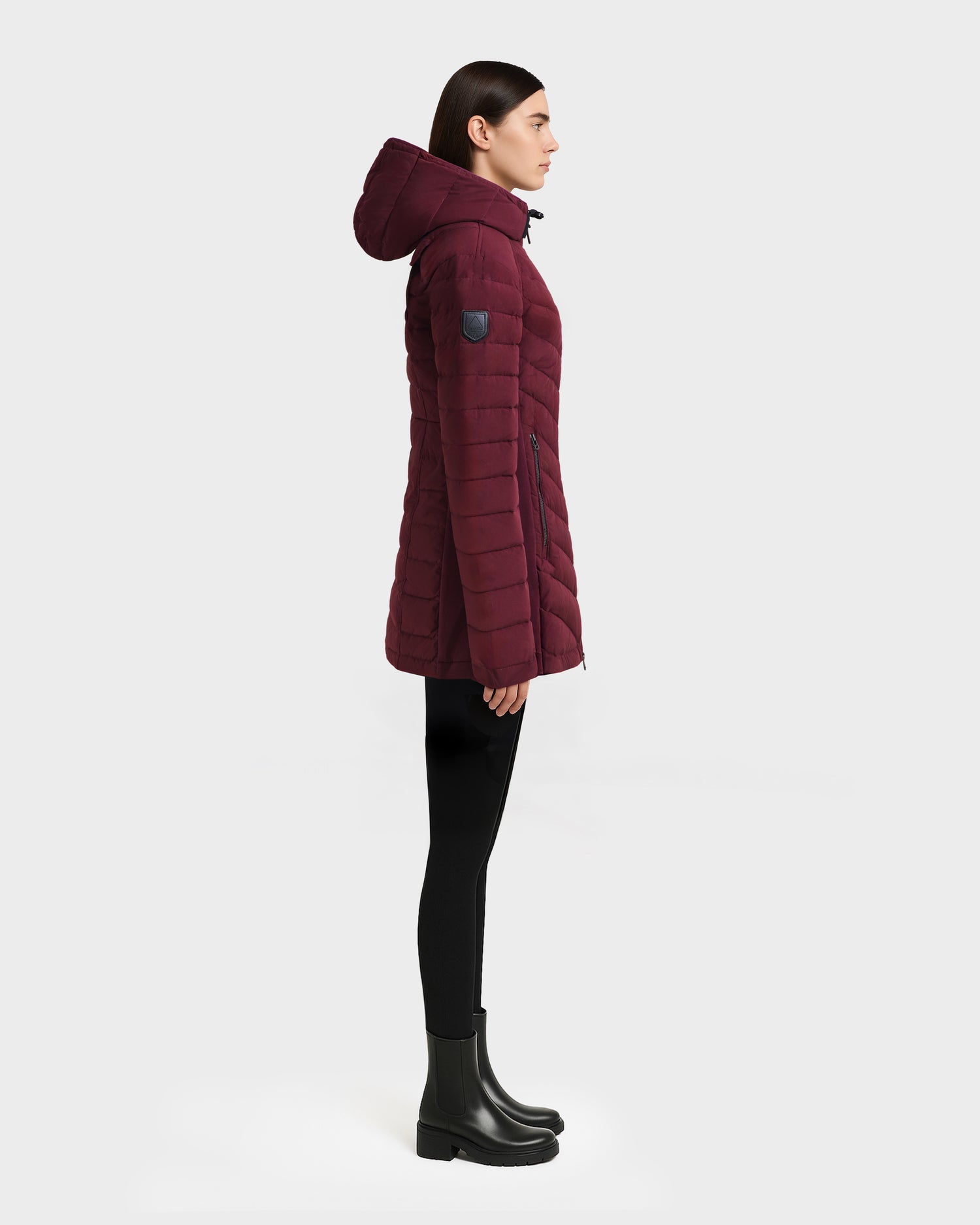 A person in profile stands against a light gray background, wearing the Rudsak CORDELIA - 4124519 Burgundy water-repellent hooded puffer jacket, paired with black leggings and black boots.