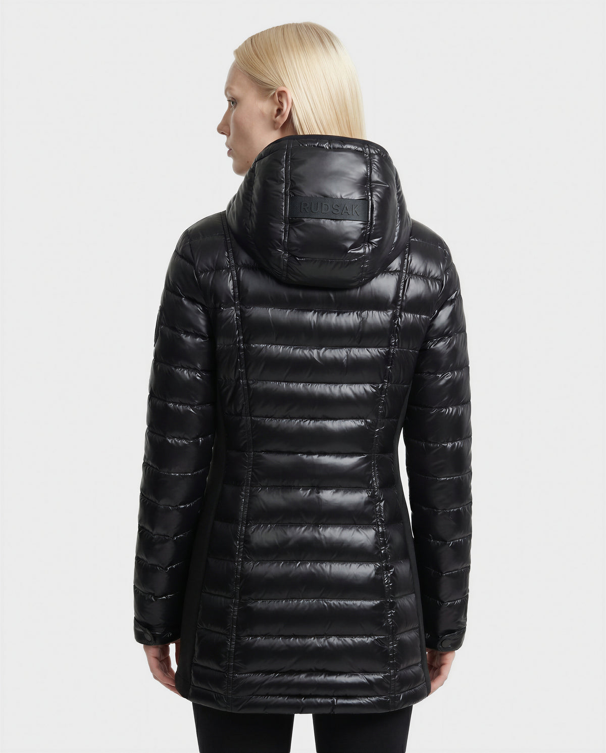 A woman with straight blonde hair, seen from behind, is wearing the Rudsak COSETTE C - L124011 Black fitted down jacket in shiny black glossy fabric with horizontal stitching and "RUDSAK" on the hood.